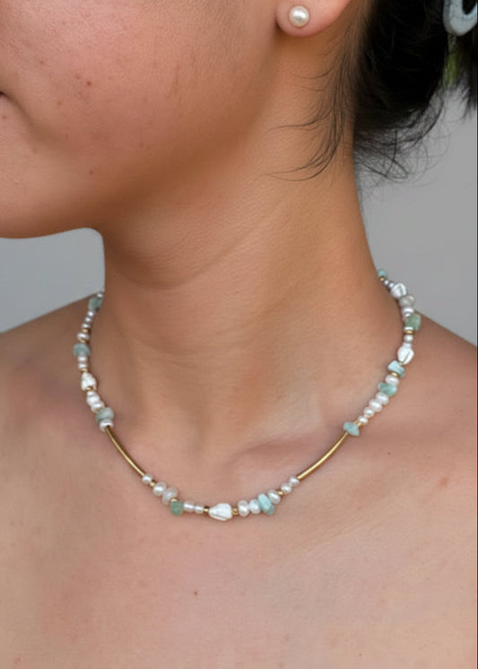 A close-up of a woman's neck and collarbone wearing a delicate necklace with white pearls, light blue beads, and a gold-toned center bar.