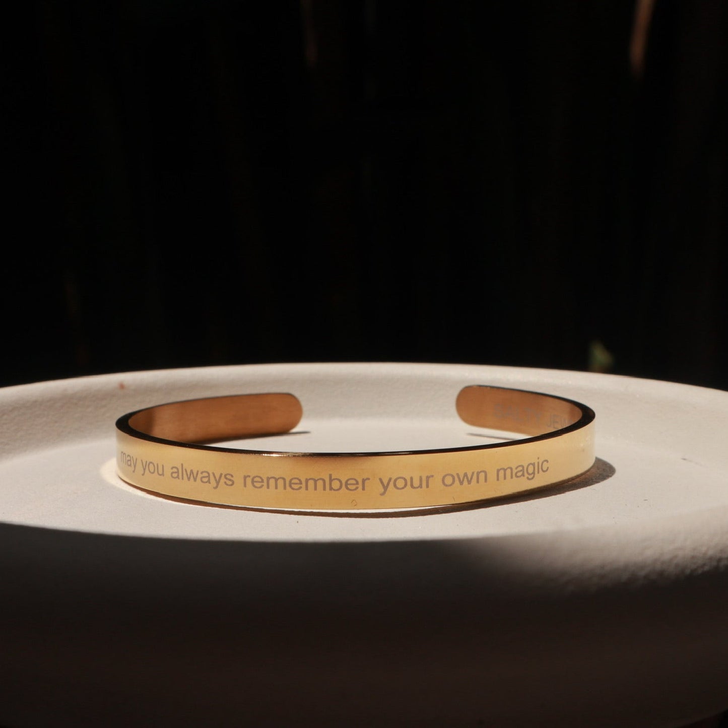 A slim gold-toned metal cuff bracelet with engraved text, displayed on a white ceramic dish against a dark background.