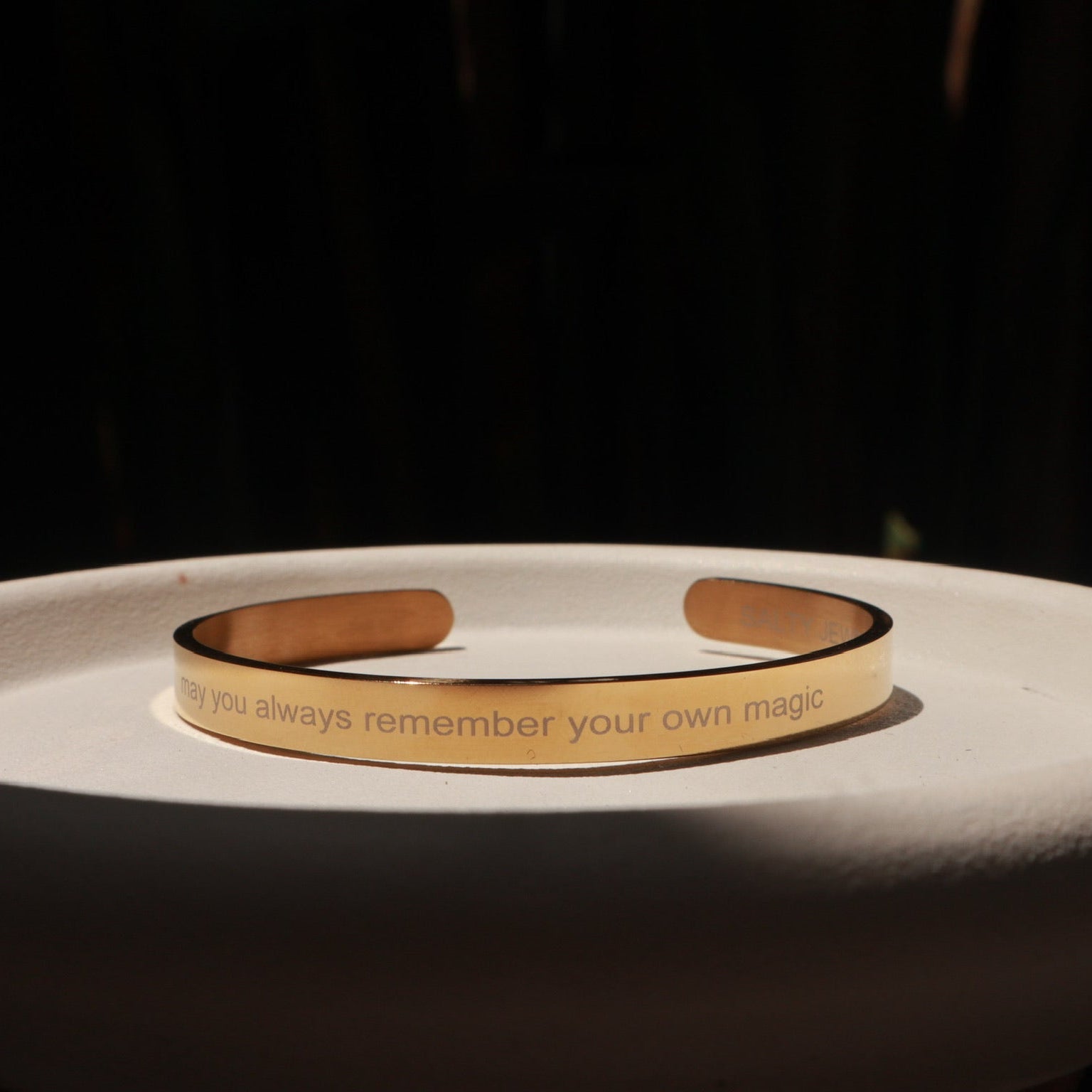 A slim gold-toned metal cuff bracelet with engraved text, displayed on a white ceramic dish against a dark background.