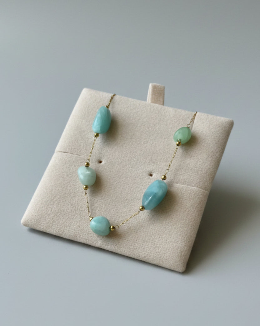 A gold chain necklace with four light blue-green stone beads displayed on a beige jewelry stand, with matching earrings in the background on a textured light blue fabric.