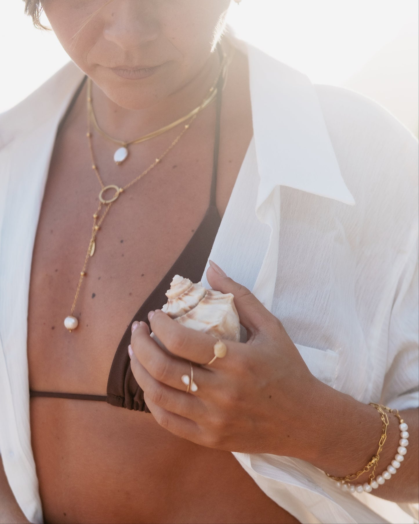 Woman holding a shell with layered necklaces and a white shirt on a bright background