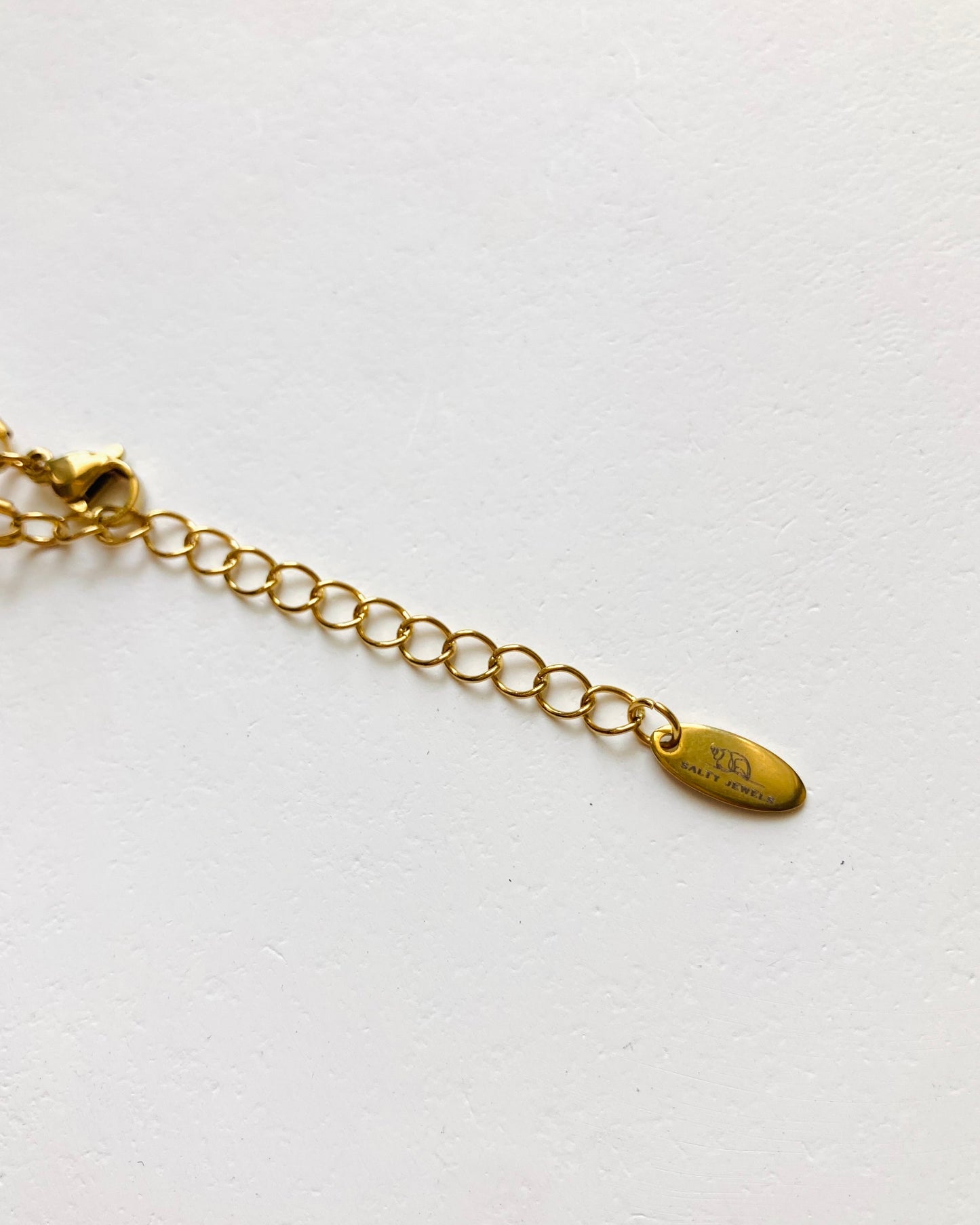 A close-up of a gold chain extender with a small oval tag engraved with 'salty jewels logo', photographed on a plain white background.