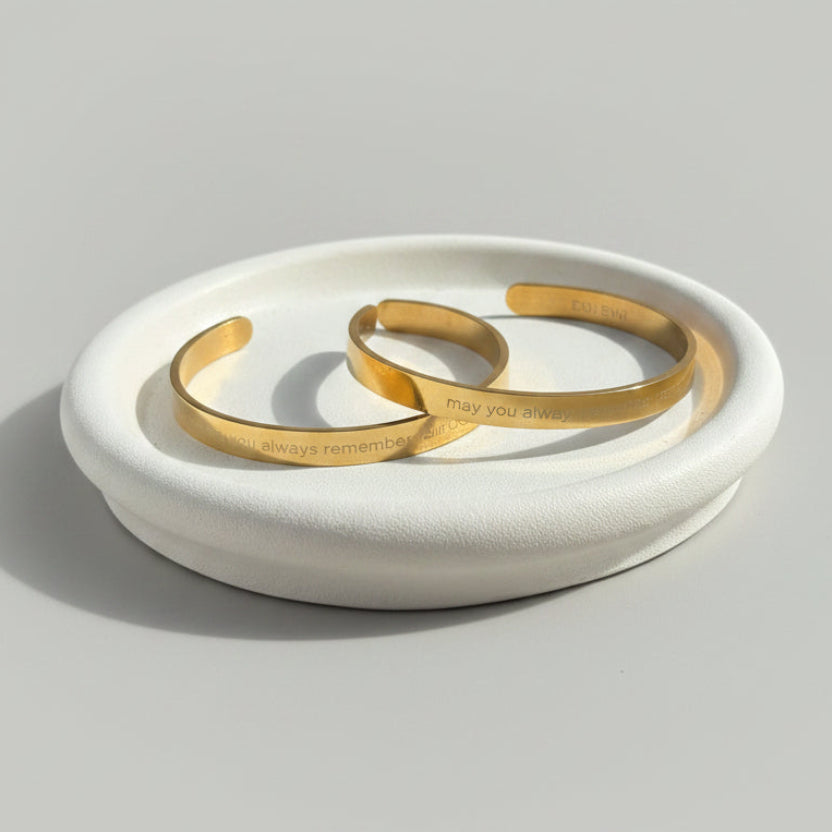 Two slim gold cuff bracelets with engraved text resting on a round white ceramic dish, set on a textured mauve tablecloth with jewelry displays and a seashell in the softly blurred background.