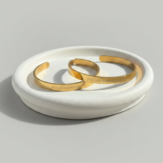 Two slim gold cuff bracelets with engraved text resting on a round white ceramic dish, set on a textured mauve tablecloth with jewelry displays and a seashell in the softly blurred background.